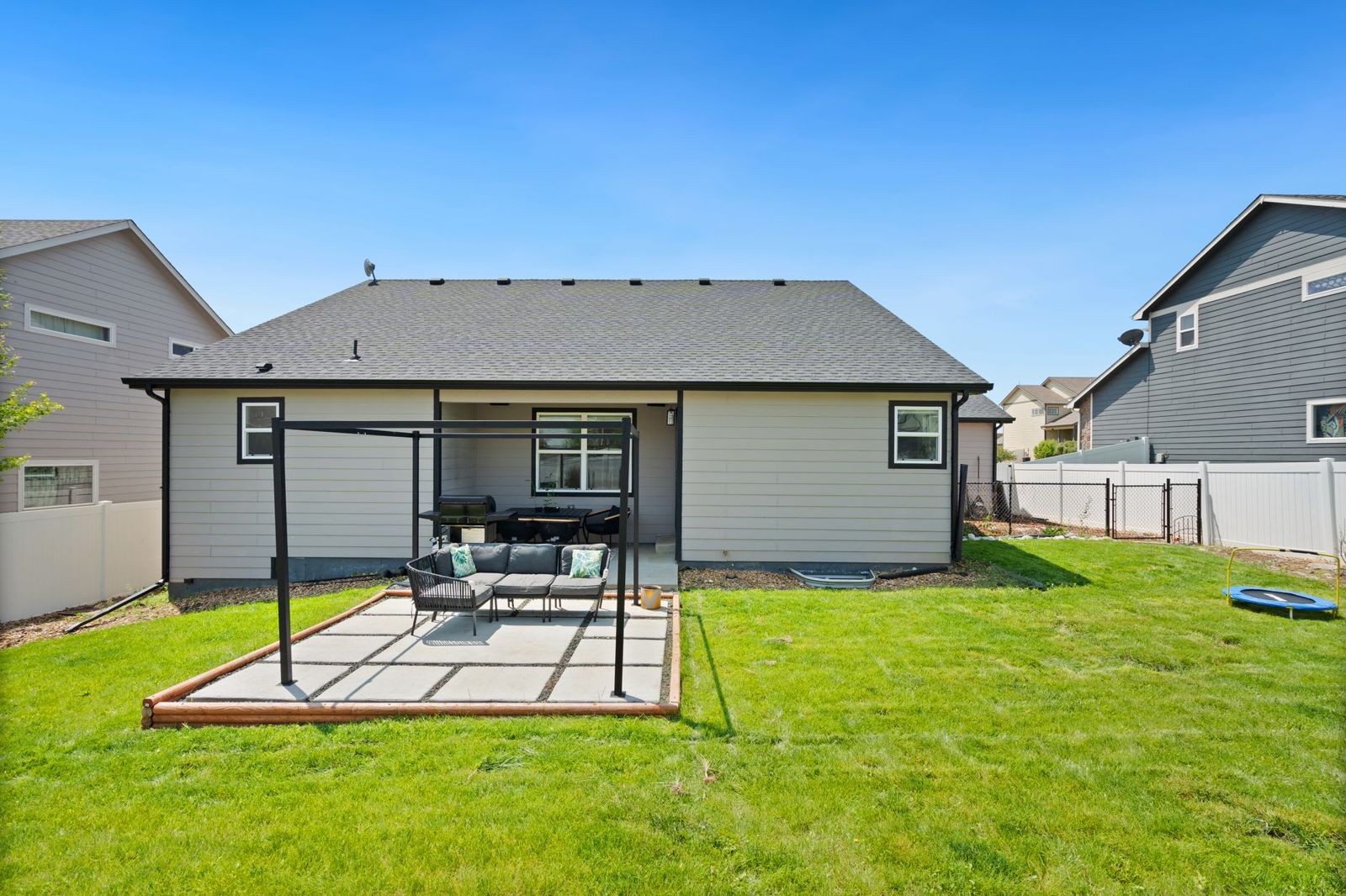 A single-story gray house with a new concrete paver patio under a black steel pergola frame, surrounded by a wide green lawn and white vinyl privacy fencing.
