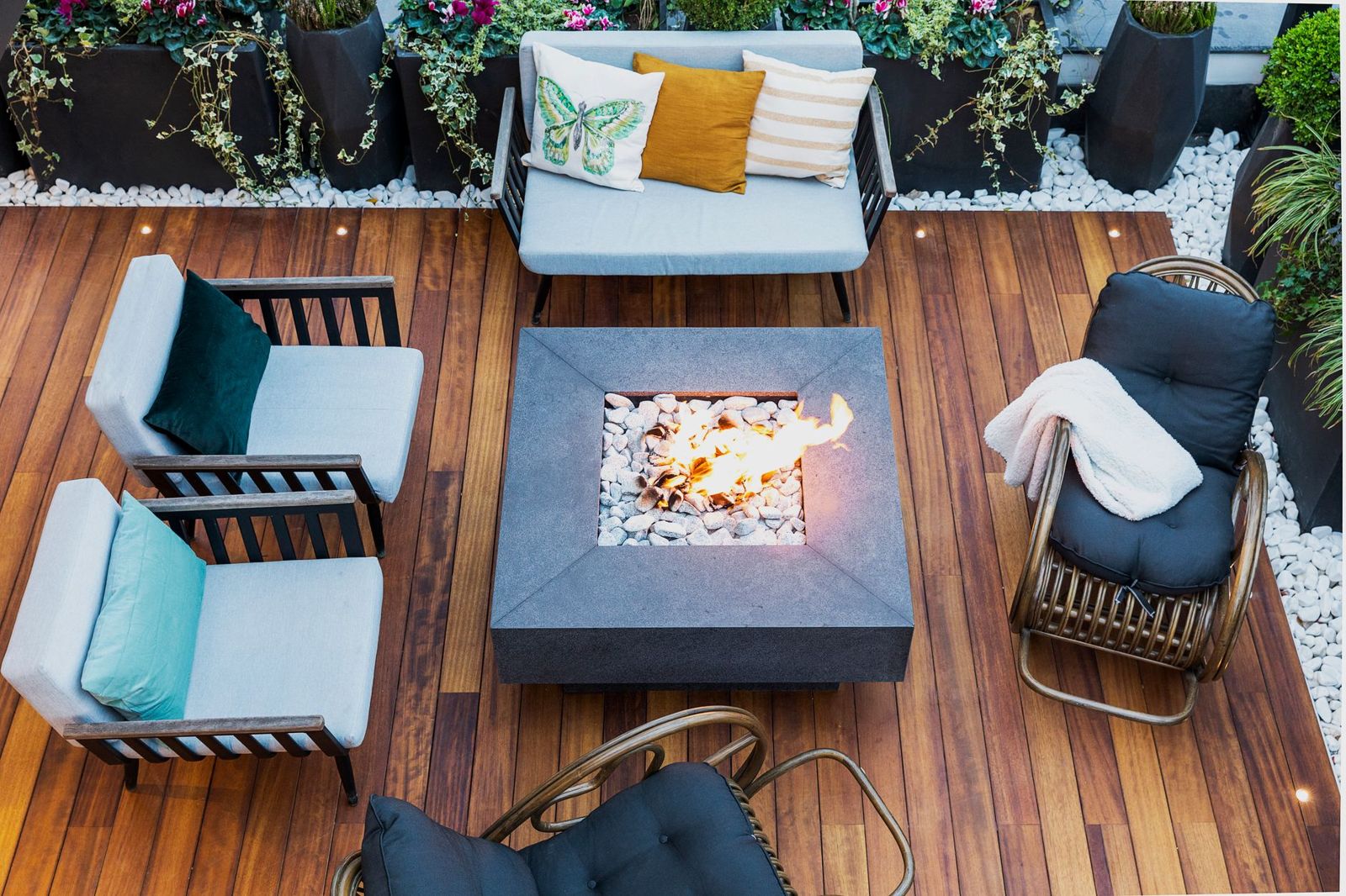 Warm-toned deck boards around a square fire table and cushioned outdoor chairs, seen from above.