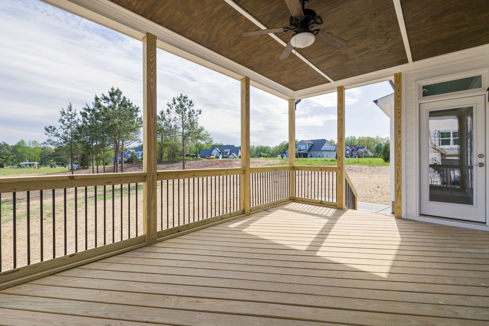 A covered porch deck made of fresh pale pressure-treated boards, with wood posts, black metal balusters, a wood-plank ceiling and a ceiling fan.