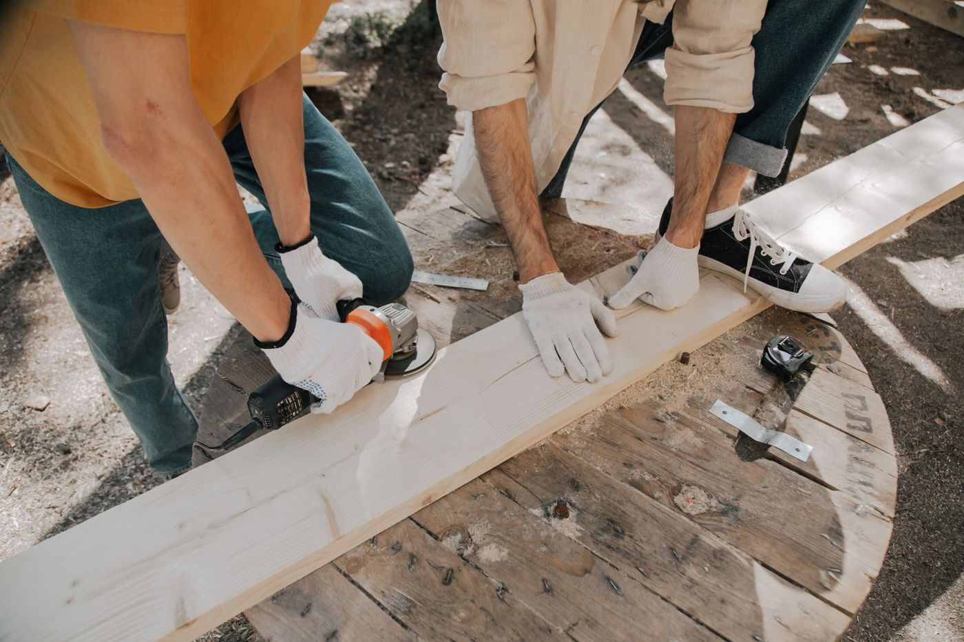 Two people in work gloves smoothing the edge of a long pale wooden board with an angle grinder on an outdoor work surface.