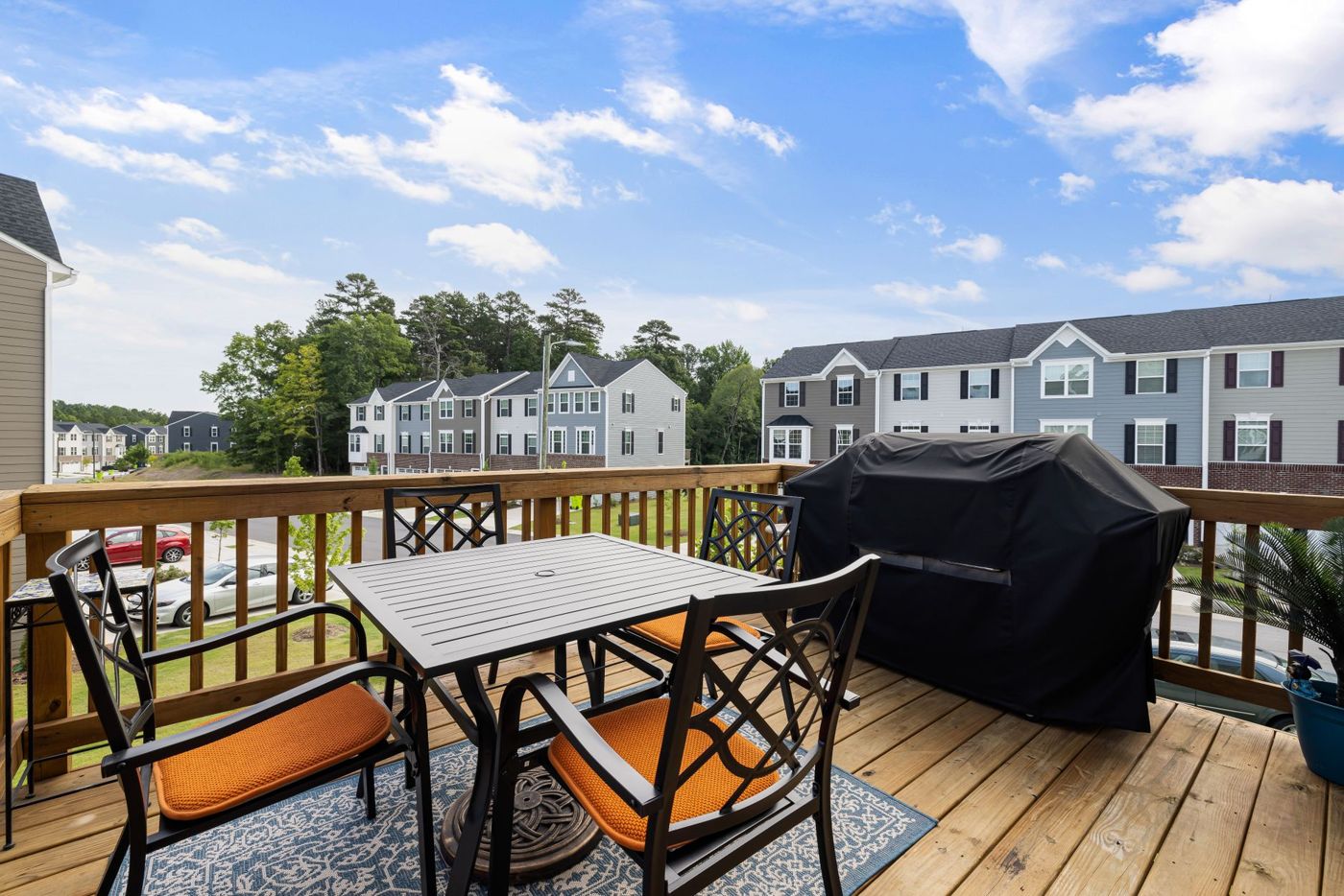 An upper-level deck with simple spindle railing, a black metal dining set with orange cushions and a covered grill, overlooking a row of townhomes.