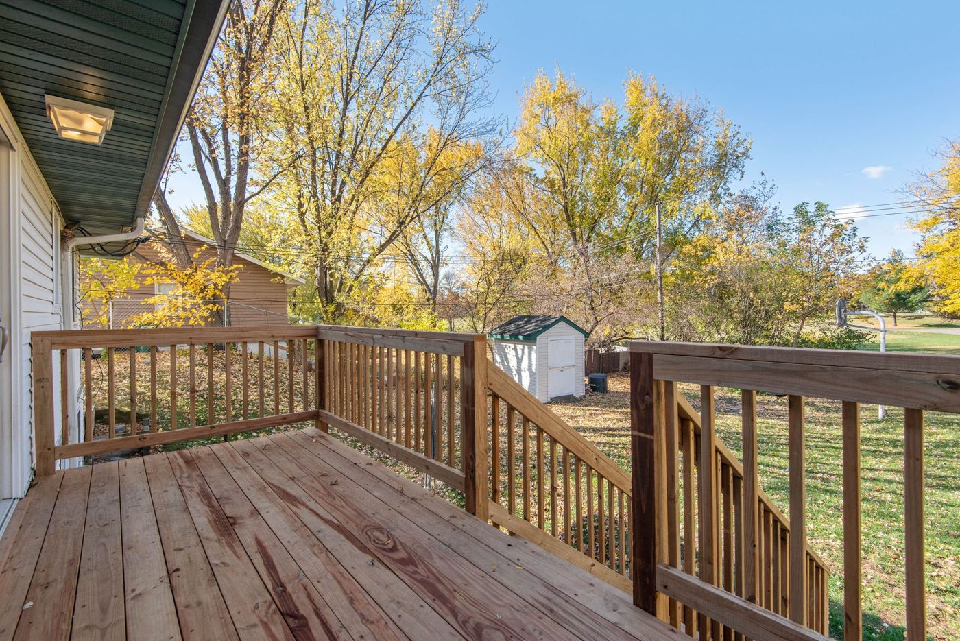 A new wooden deck off a back door with a slatted railing and stairs leading down to a leaf-covered lawn, a white shed and golden autumn trees.