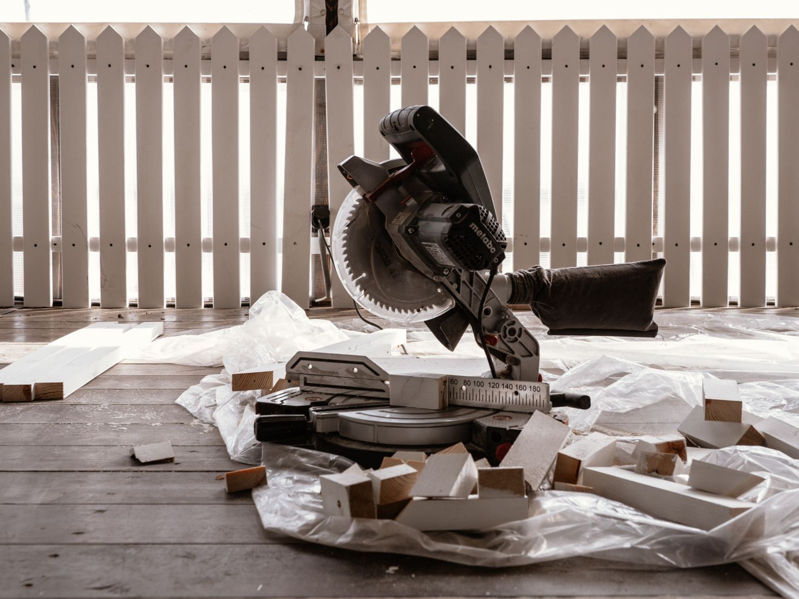 A miter saw on a weathered wooden deck covered with plastic sheeting and freshly cut white-painted wood blocks, with a white picket railing behind it.