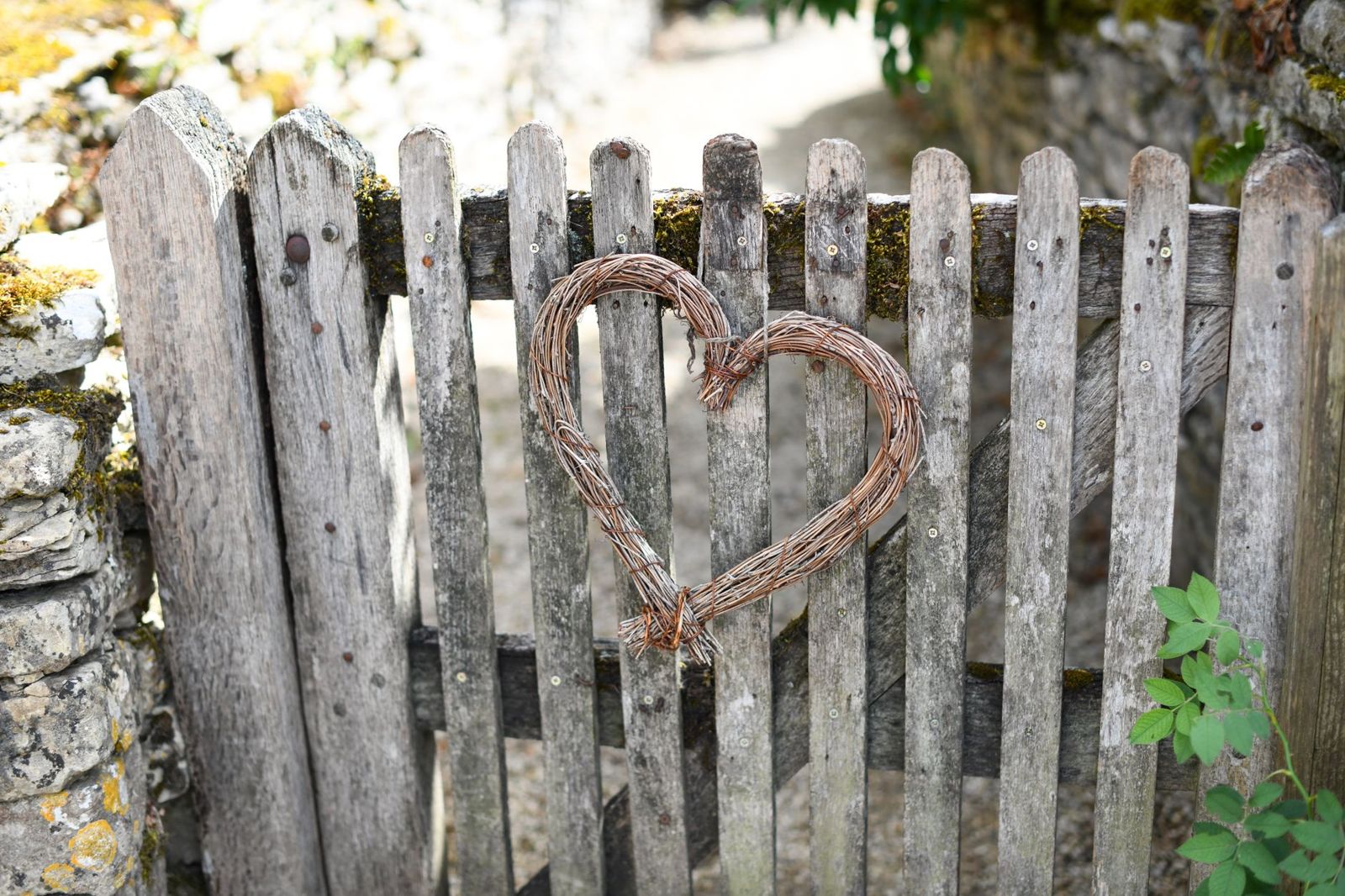 An old weathered wooden picket gate with patches of moss, a woven twig heart hung on the front, set between rough stone walls.