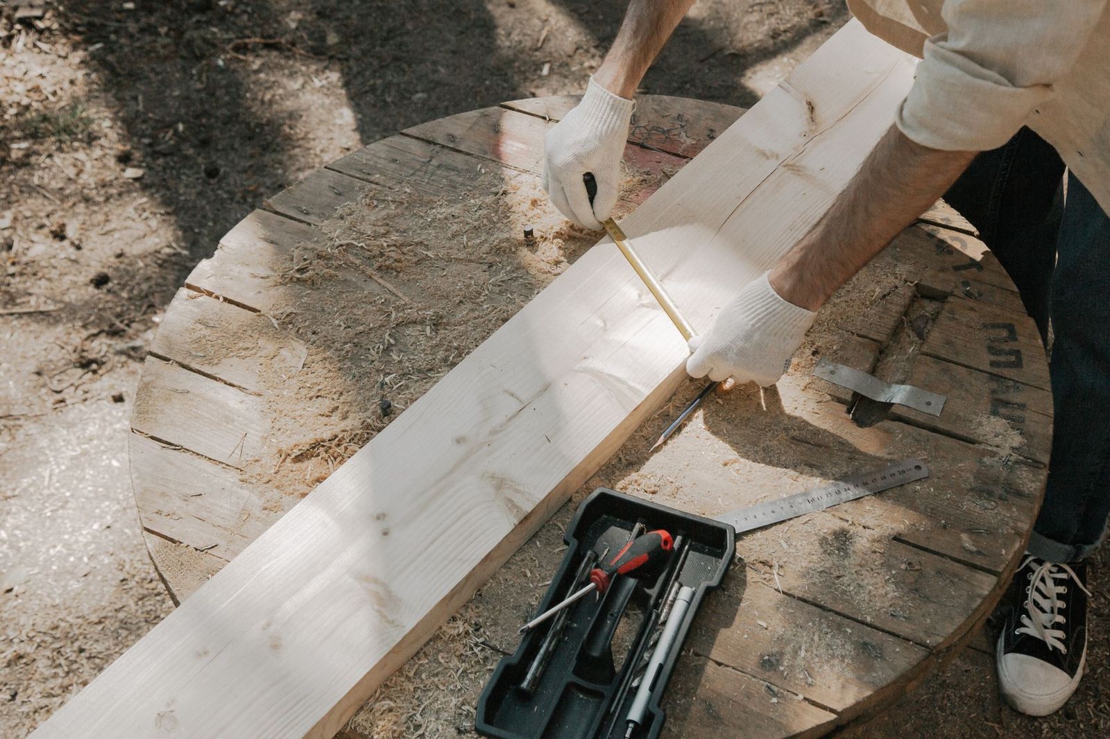 A gloved hand holds a tape measure along a pale wooden board on a round work table, with a pencil, a steel ruler and a tray of hand tools nearby.