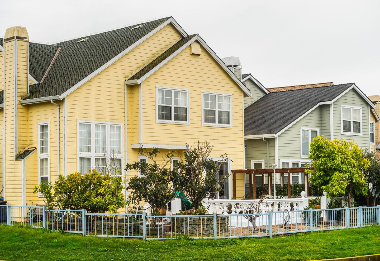 Two-story houses in yellow and sage green siding behind a low, pale blue metal picket fence and a green lawn on an overcast day.