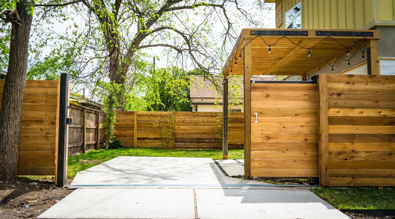 A tall horizontal cedar board fence with a matching sliding gate on a steel track, a small wood pergola with string lights, and a lawn beyond.